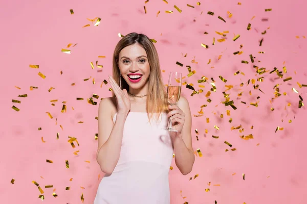 Mulher bonita com champanhe olhando para a câmera perto de confete dourado durante a festa no fundo rosa — Fotografia de Stock