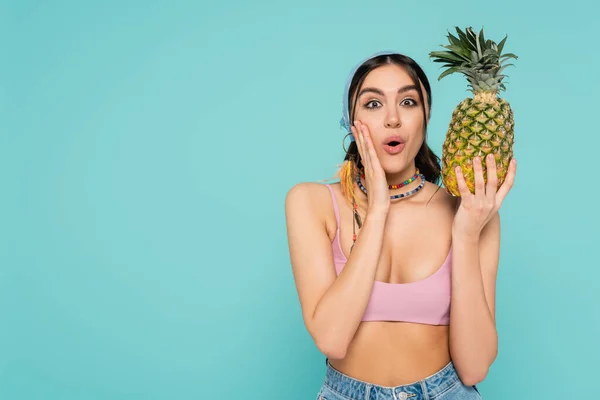 Excited woman in top and necklaces holding pineapple isolated on blue — Stock Photo