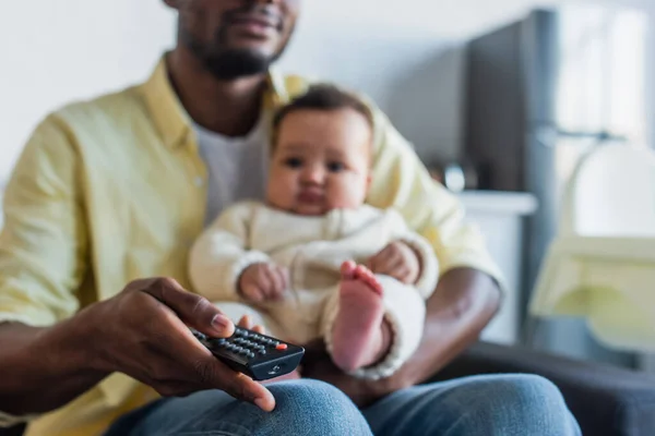 Enfoque selectivo del mando a distancia en la mano del hombre afroamericano viendo la televisión con el niño - foto de stock