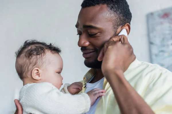 Улыбающийся африканский американец разговаривает по смартфону, держа в руках новорожденную дочь — стоковое фото
