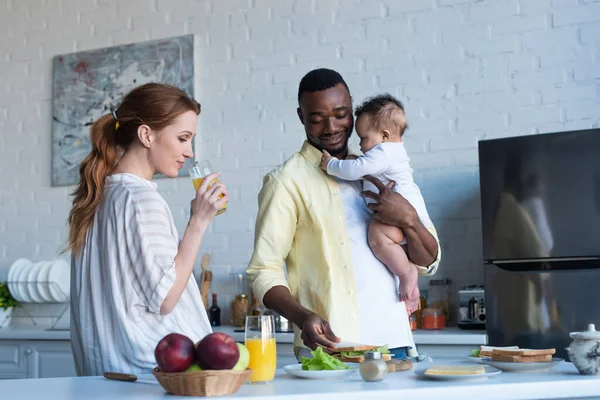 Afro-américain homme avec bébé fille préparer des sandwichs près de femme boire du jus d'orange — Photo de stock