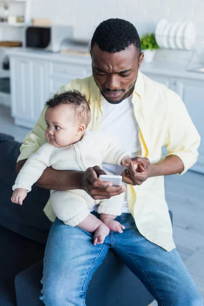 African american man sitting on couch with baby and messaging on smartphone — Stock Photo