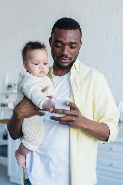 African american man holding baby girl while messaging on mobile phone — Stock Photo