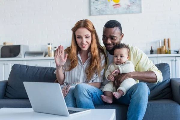 Sonriente mujer saludando mano cerca de portátil y familia interracial - foto de stock