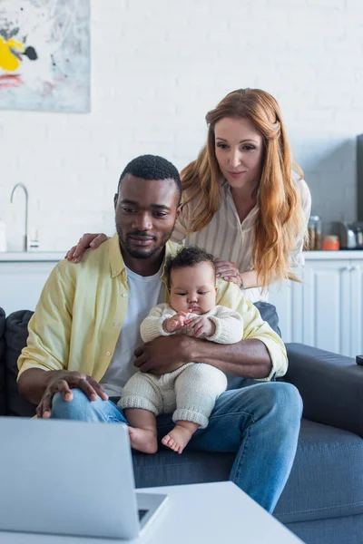 Multiethnic couple with infant daughter sitting on couch near laptop — Stock Photo