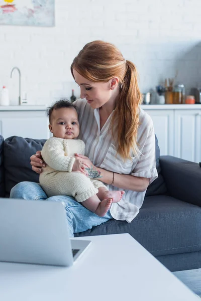 Donna seduta sul divano con neonato afro-americano figlia vicino al computer portatile — Foto stock