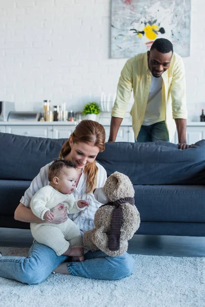 Happy african american man looking at wife playing with daughter and teddy bear on floor — Stock Photo