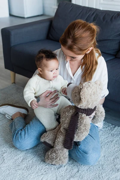 Mujer sentada en el suelo con la niña afroamericana y el oso de peluche - foto de stock