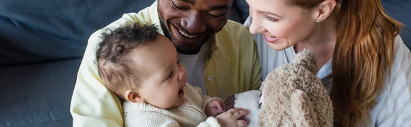Happy interracial couple looking at cheerful baby touching teddy bear, banner — Stock Photo
