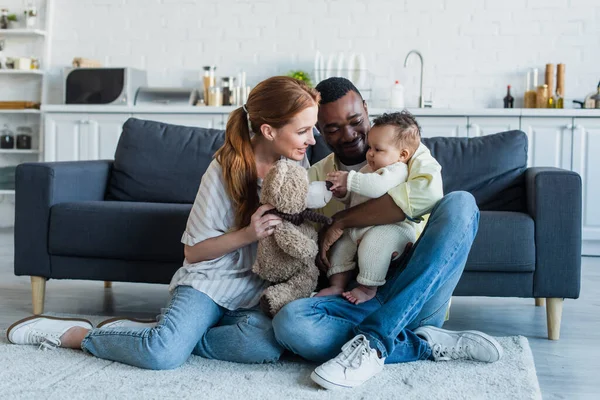 Sonrientes padres interraciales sentados en el suelo con su hija y su osito de peluche - foto de stock