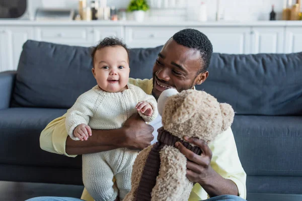 Glücklicher Vater hält fröhliches afrikanisch-amerikanisches Baby-Mädchen und Teddybär — Stockfoto
