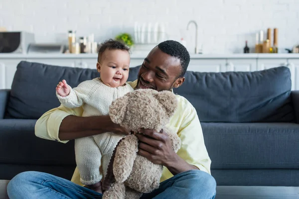 Alegre africano americano hombre sosteniendo osito de peluche mientras jugando con infantil hija - foto de stock