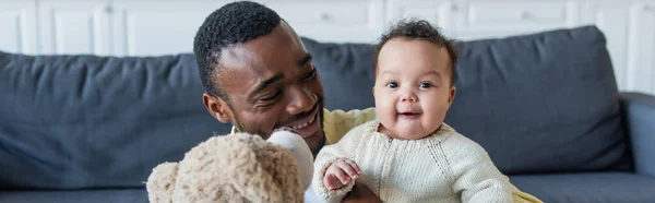 Fröhliches Baby blickt in die Kamera in der Nähe von afrikanischem amerikanischen Vater und Teddybär, Banner — Stockfoto