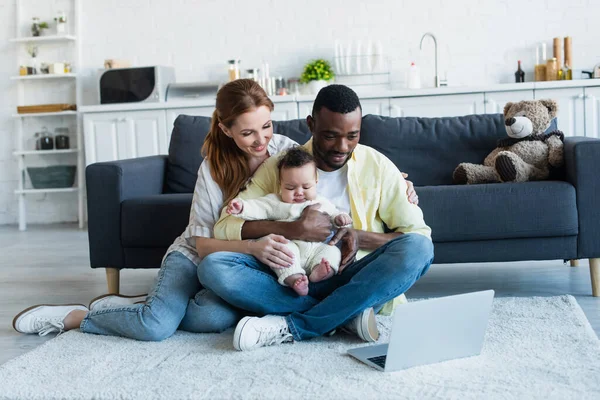 Padres multiculturales con bebé niña viendo la película en el portátil en el suelo - foto de stock