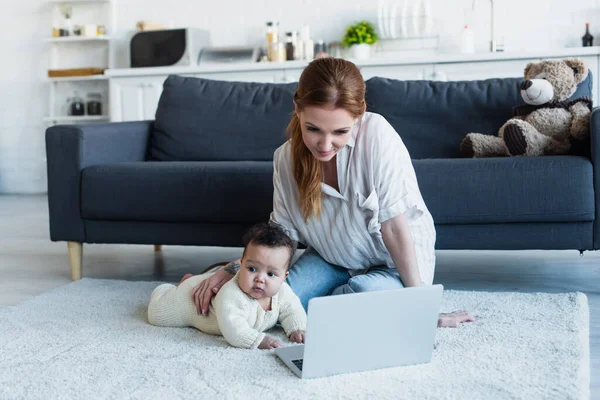 Frau schaut auf Laptop, während sie in der Nähe eines afrikanischen amerikanischen Kindes auf dem Boden sitzt — Stockfoto