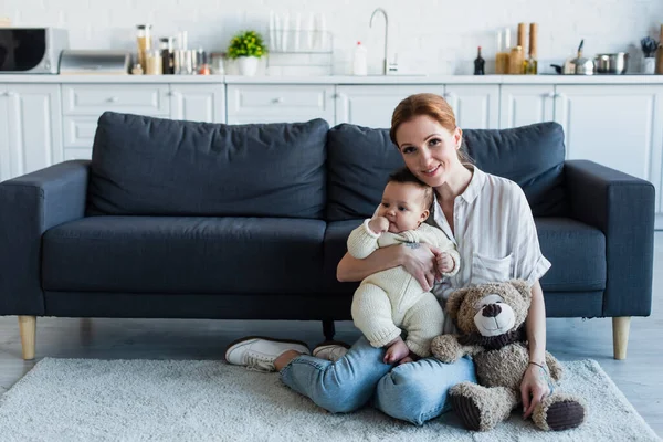 Femme heureuse souriant à la caméra tout en étant assis sur le sol avec petite fille afro-américaine près de l'ours en peluche — Photo de stock