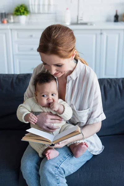 Woman reading book while sitting on sofa with infant african american kid — Stock Photo
