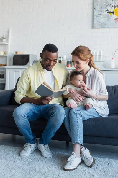 Mujer sonriente sentada en un sofá con bebé cerca del marido afroamericano leyendo libro - foto de stock