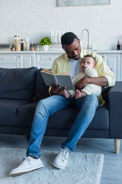 African american man sitting on sofa with baby and reading book — Stock Photo