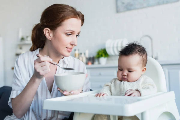 Mujer sonriente sosteniendo un tazón y una cuchara cerca de una niña afroamericana sentada en una silla de bebé - foto de stock