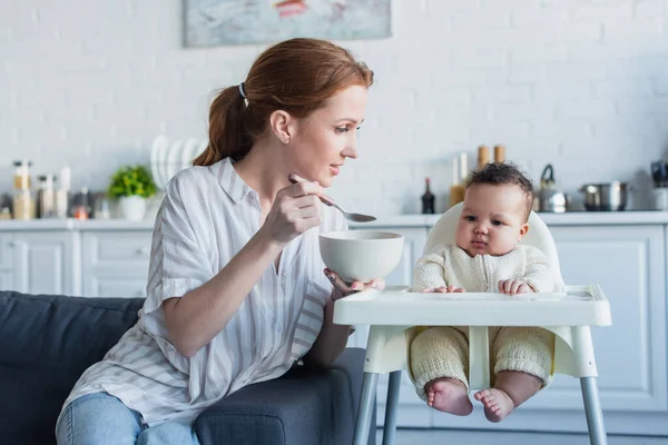 Woman feeding infant african american daughter with breakfast in kitchen — Stock Photo