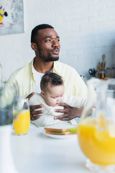Africano americano uomo guardando lontano mentre seduto con bambino vicino offuscata vaso di succo d'arancia — Foto stock