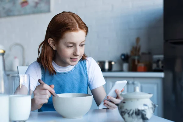 Redhead girl using smartphone while having breakfast — Stock Photo