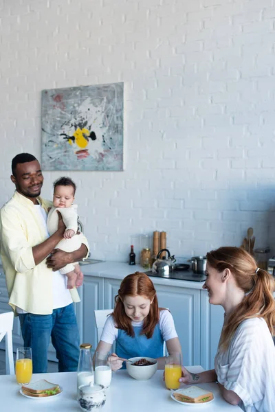 Hombre afroamericano feliz sosteniendo hija bebé cerca de la familia desayunando en cocina - foto de stock