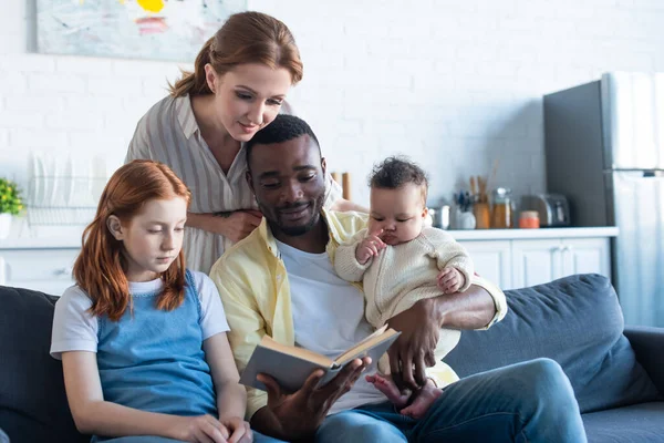 Uomo africano americano che legge libro alla famiglia multietnica sul divano di casa — Foto stock