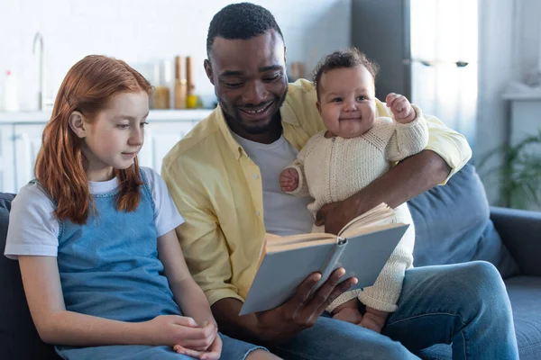 Happy african american man reading book to infant and preteen interracial daughters — Stock Photo