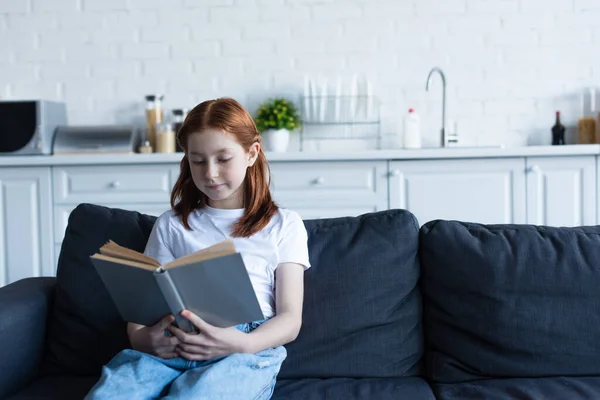 Preteen girl reading book on sofa in kitchen — Stock Photo