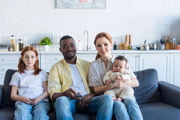 Alegre afroamericano hombre haciendo clic canales mientras mira la televisión con multiétnica familia - foto de stock