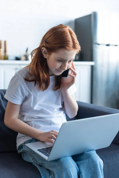 Niña preadolescente con el pelo rojo usando el ordenador portátil y hablando en el teléfono inteligente en casa - foto de stock