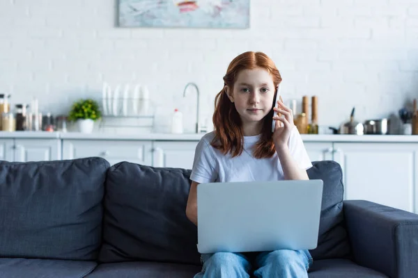 Pelirroja chica mirando a la cámara mientras habla en el teléfono móvil cerca de la computadora portátil - foto de stock