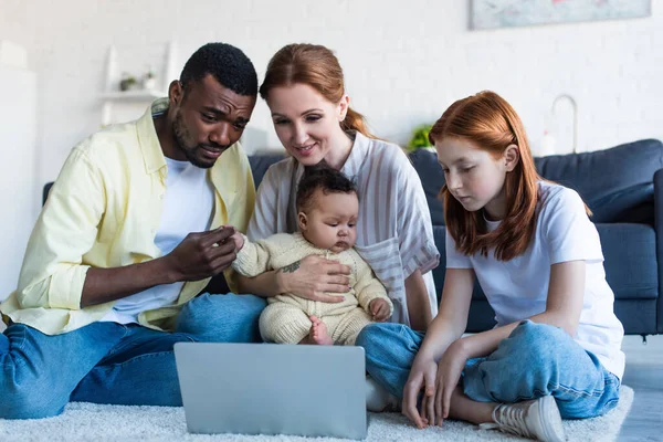 Familia interracial sentado en el suelo y viendo la película en el ordenador portátil - foto de stock