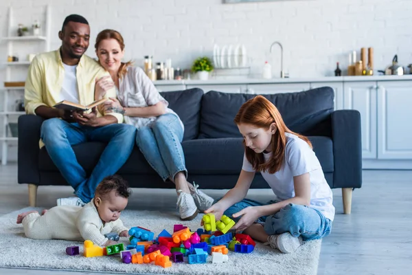 Preteen girl playing on floor with infant sister near happy interracial parents — Stock Photo
