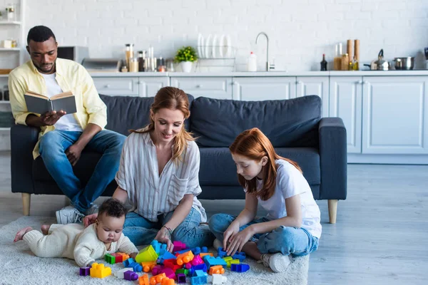 Feliz mujer jugando con interracial hijas cerca africano americano marido lectura libro en sofá - foto de stock