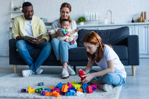 Multiethnic couple sitting on couch with infant near preteen daughter playing with building blocks — Stock Photo