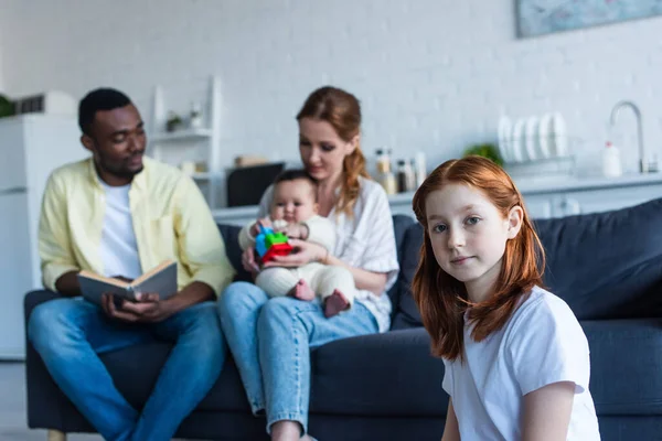 Pelirroja chica preadolescente mirando a la cámara cerca de la familia multiétnica sentado en el sofá sobre fondo borroso - foto de stock