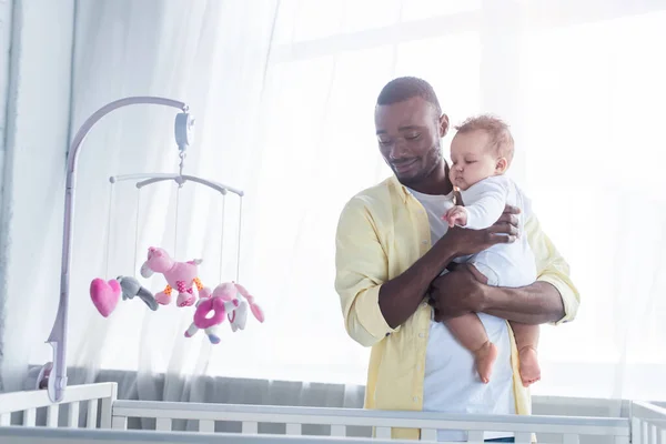 Alegre afroamericano hombre sosteniendo pequeño niño cerca cuna y bebé móvil con juguetes - foto de stock