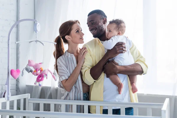 Happy interracial couple with infant daughter standing near crib — Stock Photo