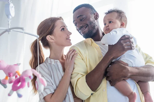 Sonriente afroamericano hombre sosteniendo bebé hija mientras mira feliz esposa - foto de stock