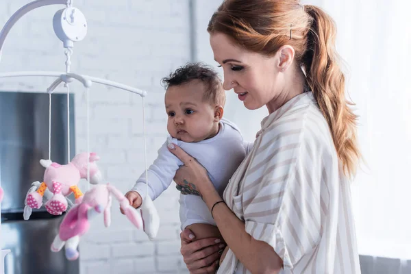 Sonriente madre sosteniendo bebé africano americano hija cerca de juguetes en bebé móvil - foto de stock