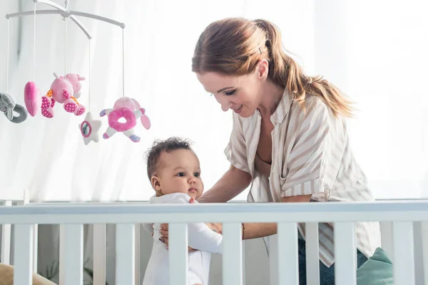 Happy woman holding african american kid near crib and baby mobile with toys — Stock Photo