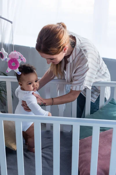 Mother supporting little african american daughter standing in crib — Stock Photo