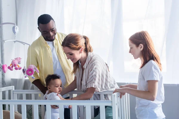 Multicultural family smiling near infant child in crib — Stock Photo