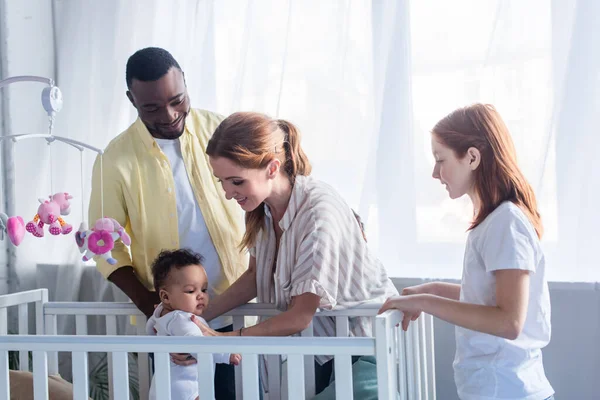 Familia multicultural sonriendo cerca del niño en la cuna - foto de stock