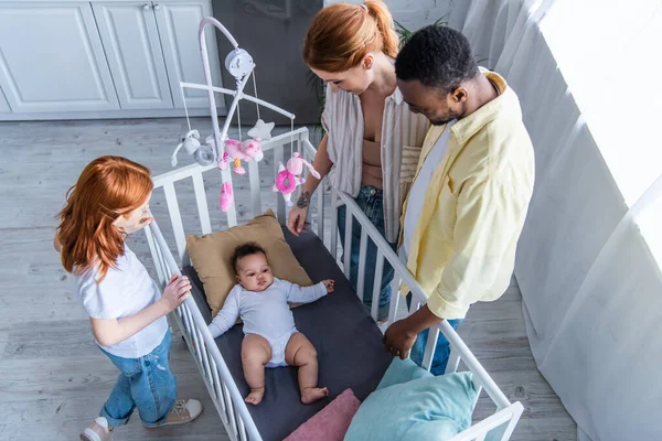 Vista aérea de la familia multiétnica mirando a la niña acostada en la cuna - foto de stock