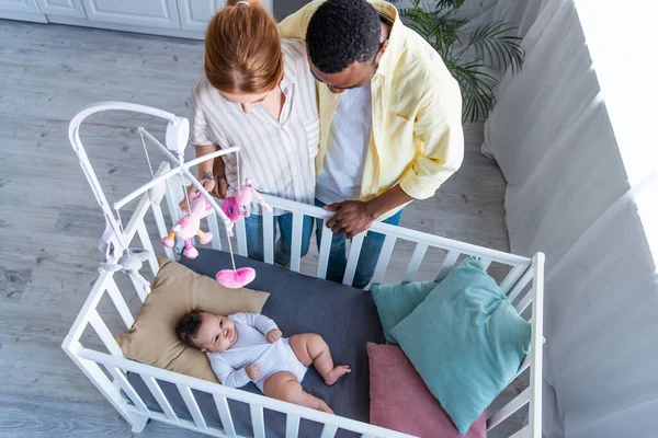 Вид сверху на межрасовую пару, стоящую рядом с ребенком, лежащим в кроватке — стоковое фото