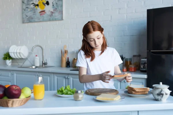 Menina ruiva preparando café da manhã com pão, queijo e alface na cozinha — Fotografia de Stock
